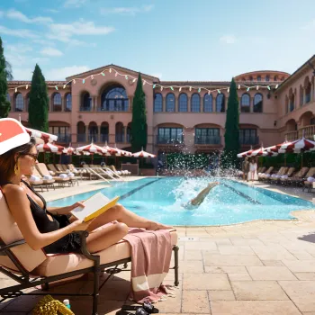 A woman wearing a Santa hat reads by a hotel pool while someone dives in, with red and white umbrellas and Mediterranean-style buildings in the background.