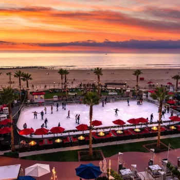 An outdoor ice skating rink by the beach at sunset, with people skating, palm trees, and seating areas with red umbrellas.