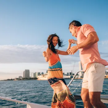 Two people dance together on the deck of a boat with the San Diego skyline and blue sky in the background.