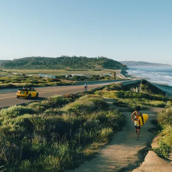 A person carries a surfboard along a path toward the beach as a yellow convertible drives on a coastal road under a clear sky.