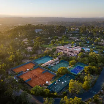 Aerial view of a sports complex with multiple tennis courts, a basketball court, nearby buildings, and lush greenery at sunset over Rancho Santa Fe in San Diego County.