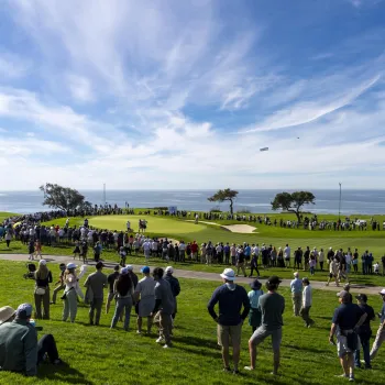 Wide shot of the Famers Insurance open with crowds watching one of the players putt on the green under blue skies and with views of the Pacific Ocean