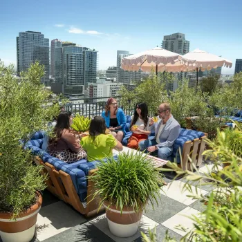 Five people sit and talk on a rooftop patio surrounded by plants, with San Diego city buildings in the background and a pink umbrella providing shade.