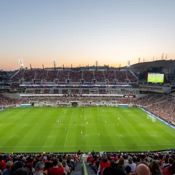 A large crowd watches a soccer match in a stadium at sunset, with both teams on the field and the scoreboard visible in the background.
