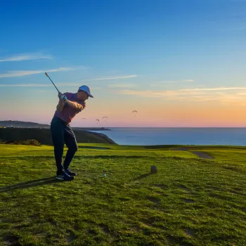 A golfer swings a club on a green golf course overlooking the ocean at sunset, with a clear sky and a few birds in the distance.