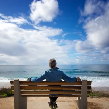 A person sits alone on a bench facing the ocean under a partly cloudy sky.
