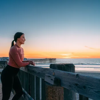 A solitary woman stands on a pier looking at a beach near sunset in San Diego.