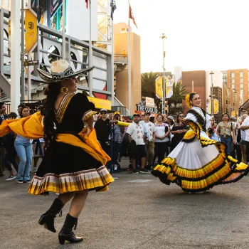 Two folklórico dancers perform in front of a crowd outside Petco Park in San Diego. The dancer in the foreground wears a black dress with yellow accents and a wide-brimmed sombrero, twirling a yellow shawl, while the dancer behind her wears a flowing white, yellow, and black ruffled dress, holding out the skirt as she spins. Baseball fans in jerseys and caps watch from behind a barrier, with stadium signage, flags, and downtown buildings visible in the background at sunset.