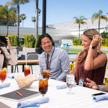 Here’s concise, accessible alt text that meets your requirements:  **Alt text:**   A small group of professionals seated around an outdoor table during a meeting at the Town & Country Resort in San Diego’s Mission Valley, with drinks, notebooks, and palm trees visible in a sunny courtyard setting.