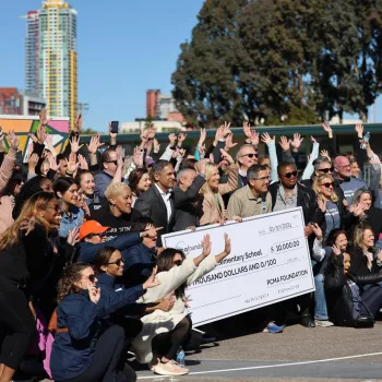 Large group of adults outdoors at a corporate volunteer or charity event, smiling and raising their hands as they pose together around two oversized donation checks held at the center of the group, with city buildings and trees in the background.