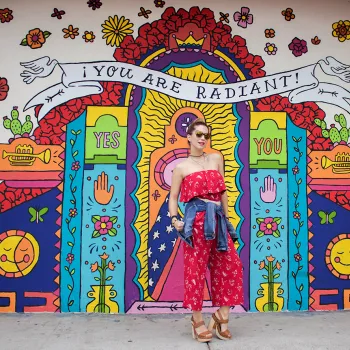 Alt text:   Colorful mural on the exterior wall of Artlexia, a Hispanic-owned business in San Diego, featuring a radiant, shrine-like design with bold, bright colors, flowers, hearts, candles, eyes, and smiling suns. A large banner at the top reads “YOU ARE RADIANT!” with “YES” and “YOU” on either side of a central glowing figure. In front of the mural, a person in a red, off-the-shoulder dress with a denim jacket tied around their waist stands on the sidewalk, looking confidently at the camera.