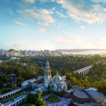 Elevated view of Balboa Park featuring the California Tower and historic buildings amid lush greenery, with the San Diego skyline stretching across the horizon and San Diego Bay beyond.
