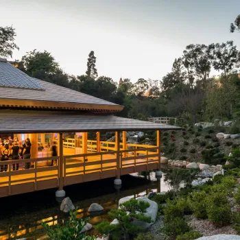 Evening view of an AANHPI cultural gathering inside a warmly lit wooden pavilion at the Japanese Friendship Garden in Balboa Park, San Diego, surrounded by stones, water, and lush hillside greenery.
