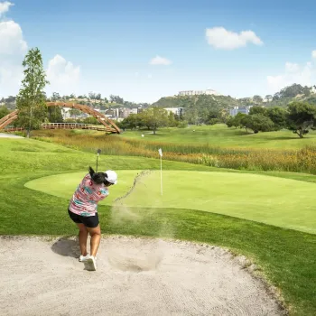 A golfer in shorts and a pink shirt drives a golf ball out of a sand trap toward the green on a sunny day in San Diego's beautiful Mission Valley.