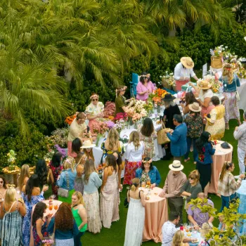 Here’s polished, descriptive alt text that includes the location you specified:  **Alt text:**   A lively outdoor event at the Estancia La Jolla Hotel & Spa in La Jolla, featuring guests gathered around pastel‑covered tables decorated with flowers, drinks, and food. Attendees in colorful, summery outfits mingle beneath tall palms and lush greenery, creating a festive garden‑party atmosphere.