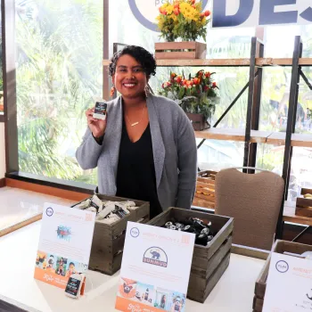 Smiling entrepreneur standing behind a display table for a Black-owned small business in San Diego, holding up a packaged product. The booth features wooden crates filled with neatly arranged bath or skincare items, promotional cards highlighting local Black-owned brands, and large windows in the background showing palm trees and a sunny Southern California landscape.