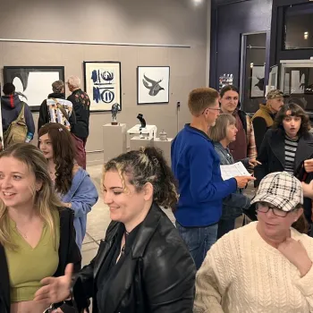 A diverse group of people mingle and chat inside The Studio Door art gallery in Hillcrest, San Diego, surrounded by colorful contemporary paintings and sculptures. Small clusters of guests sit and stand close together, smiling, talking, and looking at artwork on the walls, creating a warm, welcoming atmosphere that reflects LGBTQIA+ community event spaces and gatherings in San Diego.