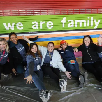 A group of seven smiling adults in matching event T‑shirts sit on a plastic-covered floor in front of a brightly colored mural in San Diego that reads ‘we are family’ with a small globe icon. They pose playfully with arms raised and legs stretched toward the camera, appearing energetic and celebratory during a community engagement event benefiting a local grade school.