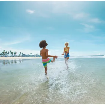 A son and his mother play in shallow water at a sunny beach, with blue skies and gentle waves.