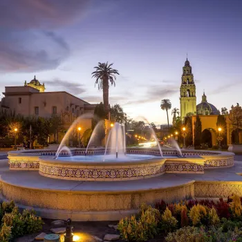 Balboa Park Fountain surrounded by vibrant flowers at dusk, with historic buildings and a clock tower in the background.