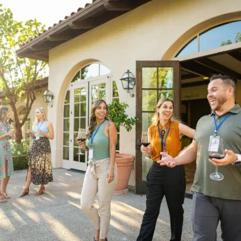 Small group networking outdoors at a San Diego venue, with attendees holding drinks and chatting near a Spanish-style building surrounded by trees and warm afternoon light.