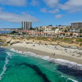 Aerial view of La Jolla Cove in San Diego, showing turquoise ocean waters, a sandy beach with visitors, rugged coastal cliffs, and nearby buildings under a bright blue sky.