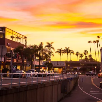 Sunset view of the Village of La Jolla in San Diego, with palm-lined streets, coastal shops and restaurants, light traffic, and a warm orange and pink sky reflecting a vibrant seaside atmosphere.