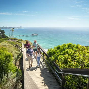 Four people in business casual attire stroll on a wooden walkway at the Scripps Coastal Meander in the La Jolla community of San Diego, against the backdrop of a sparkling blue-green sea and sunny blue skies.