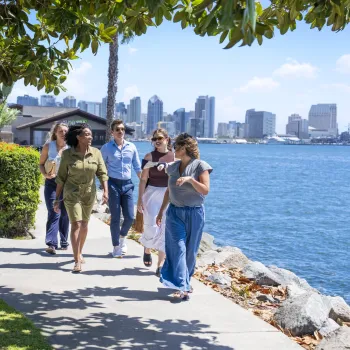 Five people walk along a waterfront sidewalk under sunny skies on Shelter Island, with the beautiful San Diego skyline in the background.