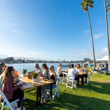 Outdoor group dining by the waterfront in San Diego, with attendees seated at tables on a grassy lawn near the bay, palm trees, and marina views under a clear blue sky.