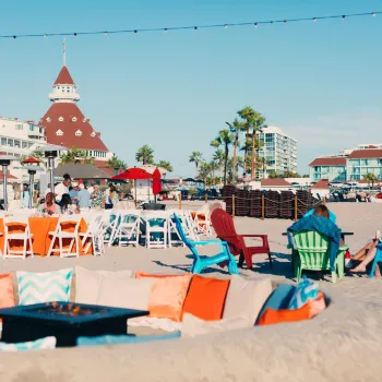 A view of the Hotel del Coronado in the beautiful San Diego region, depicting people lounging in beach chairs on the sand against a backdrop of blue skies and the hotel's brick-red spires.
