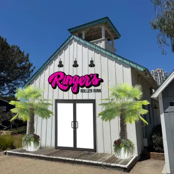Exterior of Ringer’s Seaport roller rink at Seaport Village in San Diego, featuring a white barn‑style building with the Ringer’s Roller Rink sign, palm trees in planters, and a clear blue sky.