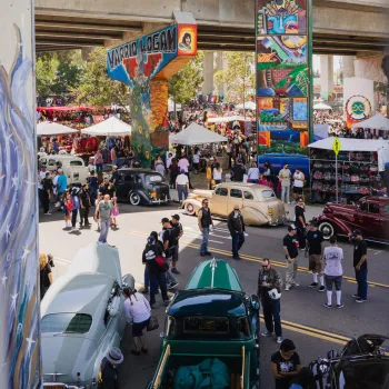 A vibrant street festival at Chicano Park in Barrio Logan under an overpass, featuring classic cars, food stalls, and colorful murals.