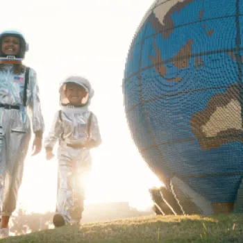 A woman and a child in astronaut suits walk past a large globe sculpture.