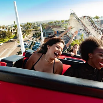 Two young women enjoy the Giant Dipper roller coaster ride at Belmont Park, laughing against a bright, sunny backdrop.