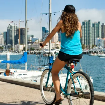 Woman riding a teal bicycle by a San Diego Bay with city buildings in the background.