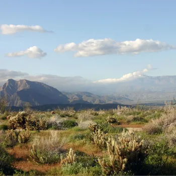 Vast desert landscape with mountains and scattered clouds under a blue sky.