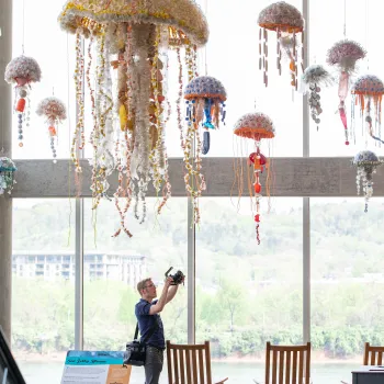A person photographing hanging jellyfish sculptures in a brightly lit room at the Washed Ashore exhibition at the San Diego Natural History Museum in Balboa Park.