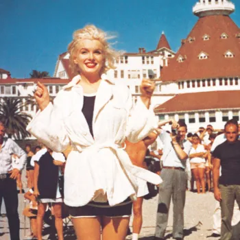 Marilyn Monroe in a white jacket stands on the beach, with a bustling crowd and the Hotel del Coronado in the background.