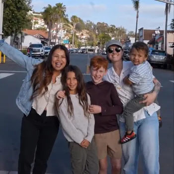 A group of five people poses joyfully under a decorative archway in a sunny street.