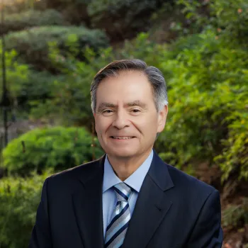 Rick Meza, CFO, in a suit stands outdoors, smiling, with greenery in the background.