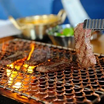 Slices of meat are being grilled over an open flame on a metal grate, with tongs holding one piece. Bowls of side dishes are visible in the background.