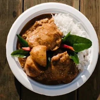 A white plate with rice, pieces of meat in brown sauce, potatoes, and green leaves, placed on a wooden surface.