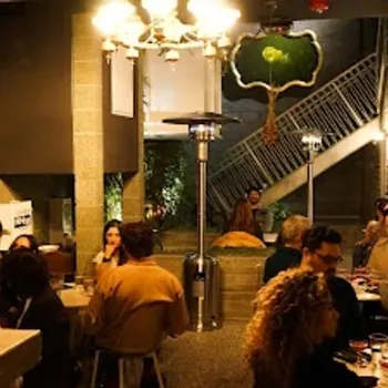 People sitting at tables in a warmly lit restaurant with a staircase and decorative tree art on the wall in the background.