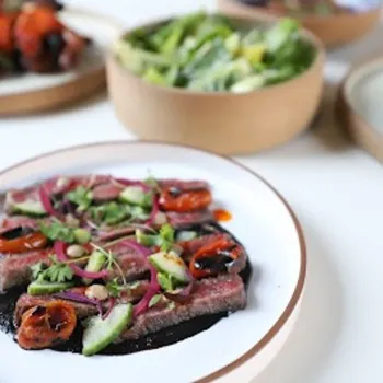 A plated dish of sliced steak topped with roasted vegetables and herbs, set on a white plate, with salads and sides in the background.