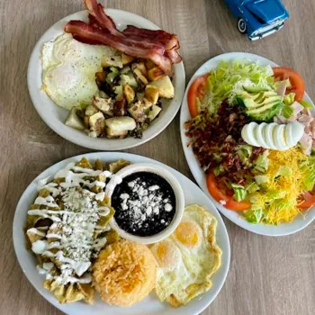 Three plates of food on a table: eggs with bacon and potatoes, a large salad with eggs and bacon, and a dish with rice, beans, eggs, and tortilla chips topped with sauce and cheese.