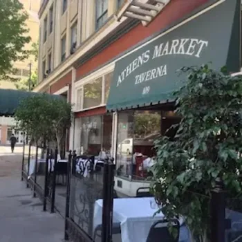 Street view of Athens Market Taverna with a green awning, outdoor seating, potted plants, and tables set for dining along the sidewalk.