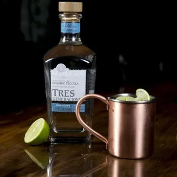 A bottle of Tres Generaciones tequila, a copper mug with lime wedges, and a halved lime are arranged on a dark wooden surface.