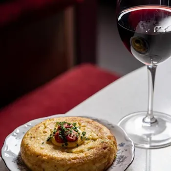 A round savory dish garnished with chopped herbs and cherry tomatoes sits on a floral plate beside a glass of red wine on a white table.