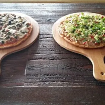 Two pizzas on wooden paddles sit side by side on a dark wooden table; the left pizza has olives, the right is topped with green vegetables.